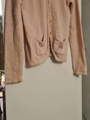 Ljusrosa stickad kofta med fickor - Söt ljusrosa stickad kofta med långa ärmar och små fickor framtill som har dekorativa knytband. Koftan har knappar längs hela framsidan och ett diskret hålmönster i materialet. Perfekt för lager på lager och en mysig look.