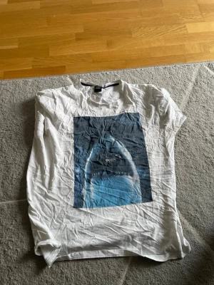 Vit t-shirt med hajtryck från BOSS - Vit t-shirt från BOSS med ett stort tryck av en haj i blå toner på framsidan. Klassisk passform och rund halsringning. Perfekt för dig som vill ha en cool och avslappnad stil. Tillverkad i mjuk bomull för skön känsla.