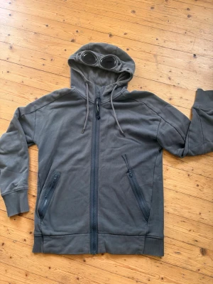 Grå hoodie med glasögon från C.P. Company - Säljer en olivgrön hoodie från C.P. Company med hel dragkedja, två fickor med dragkedja och ikoniska glasögon i huvan. Tillverkad i mjukt bomullsmaterial och har ribbade muddar vid ärmar och nederkant. 