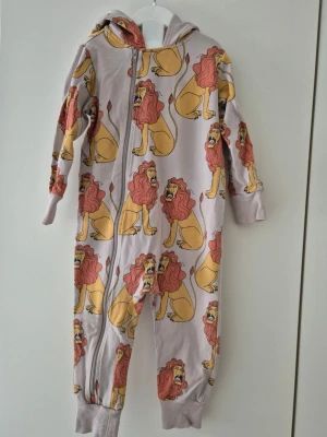 Mini Rodini lejon onesie storlek 80-86 - Säljer Mini Rodini lejon onesie med dragkedja framtill och huva. Ett eiktigt guldkorn. Storlek 80-86. Plagget har ett färgglatt lejonmönster i gult och rött över hela tyget samt ribbade muddar vid ärmar och benslut. Perfekt för dig som gillar lekfulla prints och mjuka material. Plagget är sparsamt använd trots att den är en gammal goding. Inga djur eller rökare finns i vårt hem.