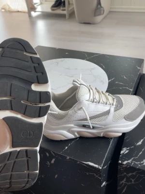 Chunky vita sneakers med grå detaljer - Snygga chunky sneakers i vitt med grå detaljer och meshpaneler för extra ventilation. Tjock sula med svart och vit färg, samt CD-logga undertill. Skorna har vita snören och en modern, sportig look som passar till streetwear.