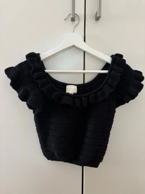 Svart stickad topp med volang - Snygg svart stickad topp från H&M med bred volang runt halsringningen och korta ärmar. Toppen har en croppad passform och är perfekt för dig som vill ha en trendig och bekväm look. Materialet är mjukt och stretchigt.