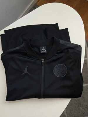 Svart PSG x Jordan träningsset - Säljer ett svart träningsset från Jordan i samarbete med Paris Saint-Germain. Setet består av en zip-jacka och matchande byxor, båda med diskreta loggor på bröstet och benet. Materialet är mjukt och stretchigt, perfekt för träning eller chill.