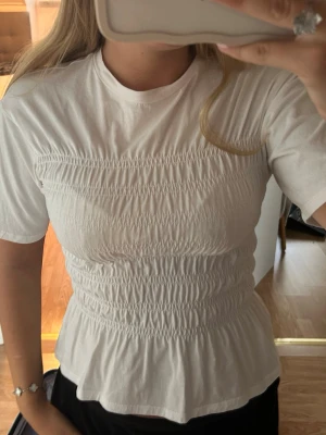 Vit smockad topp med kort ärm - Säljer en vit topp med smockad midja och bröstparti. Toppen har rund halsringning, korta ärmar och en lätt utsvängd nederkant som ger en feminin siluett. Endast använd ett fåtal gånger❤️