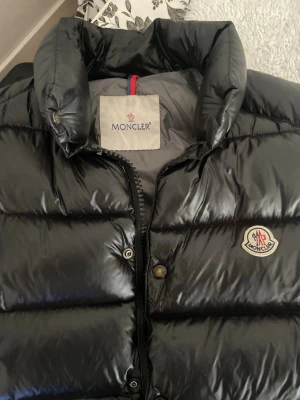 Svart dunväst från Moncler - Snygg svart dunväst från Moncler med glansig finish och klassisk logga på bröstet. Västen har hög krage, dragkedja och tryckknappar samt två fickor med dragkedja. Perfekt för lager-på-lager och streetstyle.