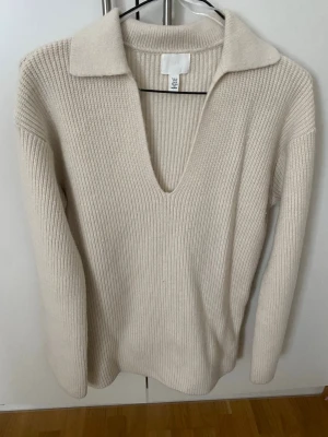 Beige ribbstickad tröja med krage - Säljer en beige ribbstickad tröja med djup V-ringning och bred krage. Tröjan har långa ärmar och är gjord i ett mjukt, tjockt material som känns riktigt cozy. Perfekt för dig som gillar en clean och stilren look.