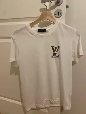 Vit  t-shirt - Snygg vit t-shirt från med klassisk  i brunt och svart på bröstet. T-shirten har rund hals och korta ärmar, perfekt för en clean och stilren look. Tillverkad i mjuk bomull för maximal komfort.