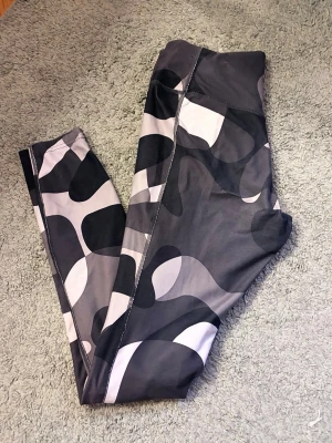 Nike Dri-FIT camo tights - Snygga träningstights från Nike med svart, grått och vitt camomönster. Tillverkade i Dri-FIT-material som transporterar bort svett och håller dig torr under träningspasset. Låg-medel midja och stretchigt tyg för bästa komfort.