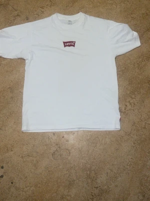Vit Levi's t-shirt med logga - Klassisk vit t-shirt från Levi's med röd logga på bröstet. Modellen är vintage fit och har rund hals och korta ärmar. Tillverkad i mjuk bomull som känns skön mot huden. Perfekt till jeans eller shorts för en clean och enkel stil.