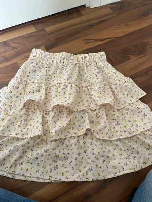 Ljus volangkjol med småblommigt mönster - Supersöt ljus kjol med volanger och småblommigt mönster i lila och gult. Kjolen har resår i midjan och är gjord i ett lätt, flowy tyg som ger en romantisk känsla. Perfekt för vår och sommar!