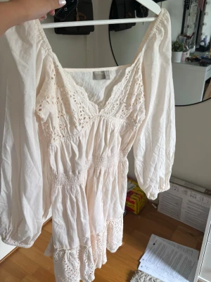Vit kort klänning med broderi - Söt vit kortklänning med broderade detaljer och v-ringning. Klänningen har långa ärmar och smock i midjan för en bekväm passform. Tillverkad i lätt bomull, perfekt för varma dagar. Romantisk och luftig stil med volangkant nertill.