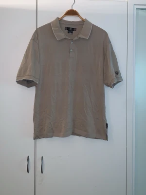 Beige pikétröja från Nike Golf - Snygg beige pikétröja från Nike Golf med klassisk krage och tre knappar framtill. Tröjan har korta ärmar och diskreta vita detaljer vid kragen. Perfekt för en sportig och avslappnad stil.