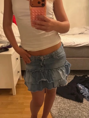 Blå volangkjol i jeans från Gina Tricot - Säljer en blå jeanskjol från Gina Tricot med volangdetaljer och råa kanter. Kjolen har en kort modell och är perfekt för dig som gillar en avslappnad men trendig stil. Passar till både sneakers och boots.