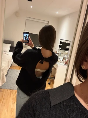 Svart stickad tröja med öppen rygg - Trendig svart stickad tröja med öppen rygg och knytband i nacken. Tröjan har en lös passform, långa ärmar och är gjord i ett mjukt, fluffigt material. Perfekt för dig som vill ha en unik twist på din vardagslook.