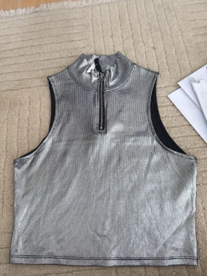 Silvrig croppad polotopp med dragkedja - Snygg silvrig croppad polotopp i syntetmaterial med half zip-dragkedja framtill. Ärmlös design och metallisk finish som ger en futuristisk vibe. Perfekt för dig som vill sticka ut med en trendig topp.