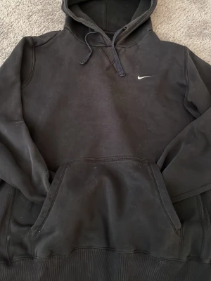 Svart hoodie från Nike med huva - Svart hoodie från Nike med klassisk huva och dragsnören. Liten vit Nike-logga på bröstet och stor magficka framtill. Tillverkad i mjukt material, perfekt för en avslappnad stil. Långa ärmar och ribbade muddar.