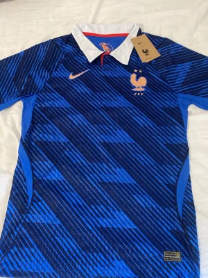 Blå Frankrike fotbollströja Nike - Snygg blå fotbollströja från Nike med Frankrikes landslagsemblem och två stjärnor på bröstet. Tröjan har ett diagonalrandigt mönster i olika blå nyanser, vit krage och orange Nike-logga. Tillverkad i lätt och ventilerande material, perfekt för träning eller match.