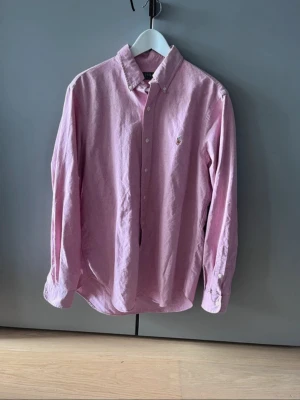 Rosa skjorta från Polo Ralph Lauren - Klassisk rosa skjorta från Polo Ralph Lauren med broderad logga på bröstet. Skjortan har knappar framtill, button-down krage och långa ärmar. Perfekt för en clean och stilren look.