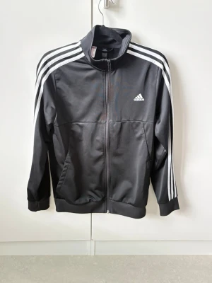 Svart Adidas träningsjacka - Snygg svart träningsjacka från Adidas med dragkedja framtill och klassiska vita ränder längs ärmarna. Jackan har hög krage, två sidofickor och Adidas-logga på bröstet. Tillverkad i ett lätt och funktionellt material, perfekt för träning eller chill.