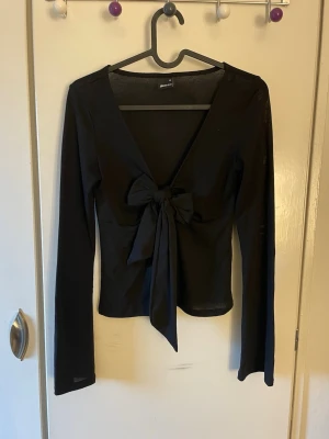 Svart långärmad topp med knytning - Svart tunn långärmad topp med v-ringning och slits med knytning framtill. Toppen har en croppad passform och är tillverkad i ett mjukt, stretchigt material som sitter bekvämt på kroppen. Från Gina tricot i storlek XS. 