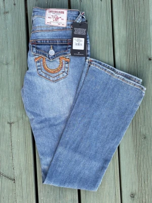 Blå True Religion jeans bootcut - Snygga blå jeans från True Religion med klassisk bootcut-modell. Jeansen har dekorativa sömmar i orange och gult på bakfickorna samt logga på bakre linningen. Tillverkade i denim med en autentisk tvättad look och detaljerade fickor.
