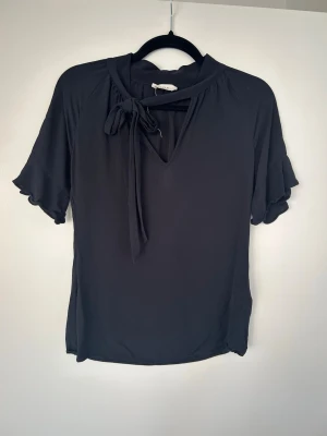 Svart blus med knytband - Snygg svart blus med korta volangärmar och dekorativt knytband vid halsen. Blusen har en lätt v-ringning och är tillverkad i ett mjukt, tunt tyg som faller fint. Perfekt för en trendig och stilren look.