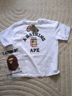 Vit A Bathing Ape x Burberry t-shirt - Säljer en vit t-shirt från A Bathing Ape i storlek XXL. T-shirten har klassiskt tryck med BAPE-loggan och texten på bröstet, samt coola camo-detaljer på taggen. Perfekt för dig som gillar streetwear och vill sticka ut.