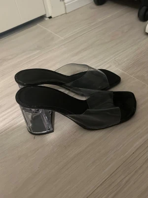 Svarta pumps med transparent klack - Säljer ett par trendiga svarta pumps med bred transparent klack och genomskinlig rem över foten. Skorna har öppen tå och en chunky klack i plast som ger en cool look. Perfekta för dig som vill sticka ut med en modern och stilren sko.