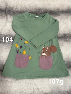 Grön barntunika med ekorre och fickor - Grön långärmad barntunika med två lila fickor framtill. Klänningen har ett motiv av en ekorre och färgglada blommor. bekväm modell med rund halsringning, perfekt för lek och bus.🌸 Fint använt skick. Storlek 104