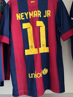 Barcelona Neymar Jr matchtröja 20014/15 - Säljer en FC Barcelona matchtröja med Neymar Jr och nummer 11 på ryggen. Tröjan har blå och röda vertikala ränder, klubbmärke på bröstet och Qatar Airways som sponsor. Tillverkad i lätt och ventilerande material, perfekt för fotboll eller träning.