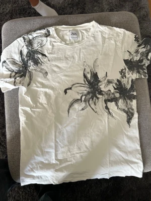 Vit t-shirt med svart blommönster Zara - Snygg vit t-shirt från Zara med svart blommigt mönster över axlar och bröst. Klassisk rund halsringning och korta ärmar. Tillverkad i mjuk bomull som känns skön mot huden. Perfekt för dig som vill ha en unik och stilren look.