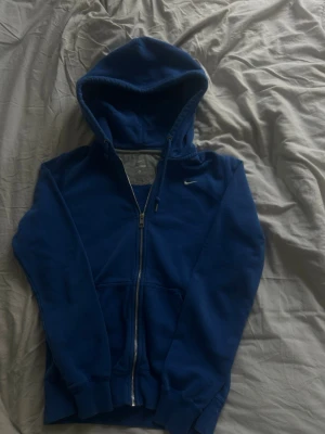 Blå zip hoodie från Nike - Blå hoodie från Nike med dragkedja framtill och klassisk huva. Tröjan har fickor på magen och ribbade muddar vid ärmslut och nederkant. Insidan är mjukt fodrad i grått för extra komfort. Perfekt för en avslappnad streetwear-look.