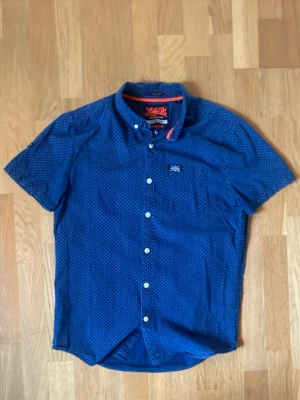 Superdry - Kortärmad skjorta | Marinblå mönster | Herr | Strl L - Snygg blå skjorta från Superdry med vita prickar och korta ärmar. Skjortan har klassisk krage, bröstficka med Superdry-logga och vita knappar framtill. Tillverkad i mjuk bomull och har en regular fit. Perfekt för en avslappnad men stilren look. Mycket bra använt skick! Jag är öppen för bud😊