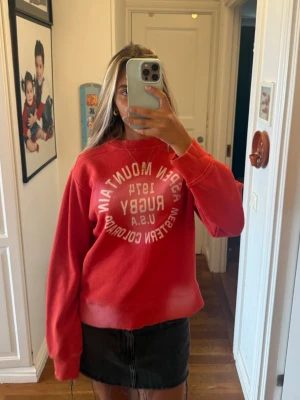 Röd sweatshirt med tryck - Cool röd sweatshirt med vit text på både fram- och baksidan. Texten säger 'Aspen Mountain 1974 Rugby U.S.A Western Colorado'. Tröjan har avslappnad passform, rund halsringning och ribbade muddar. Perfekt till jeans eller kjol för en casual look.