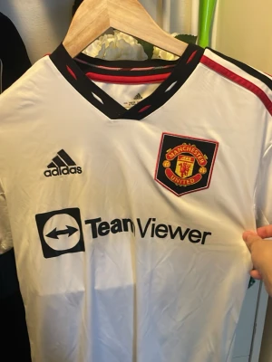 Manchester United bortatröja Adidas - Snygg vit bortatröja från Manchester United med svart och röd detalj på kragen och axlarna. Broderat klubbmärke på bröstet och Adidas-logga. Tillverkad i lätt och ventilerande material, perfekt för fotboll eller träning.