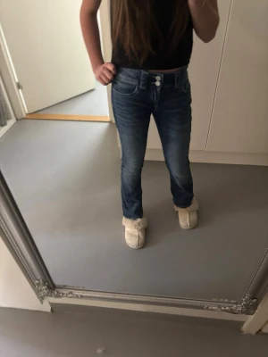 Blå bootcut jeans med fickdetaljer - Säljer ett par blå bootcut jeans med snygg tvätt och dubbla knappar i midjan. Jeansen har två fickor bak med lock och knapp, samt klassiska framfickor. Perfekta till sneakers eller boots och ger en avslappnad men trendig look.