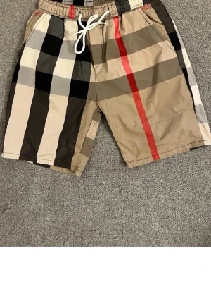 Rutig shorts med snörning - Snygga shorts med klassiskt rutigt mönster i beige, svart, vitt och rött. Shortsen har elastisk midja med vit snörning och sidofickor. Perfekta för sommaren och ger en trendig look.