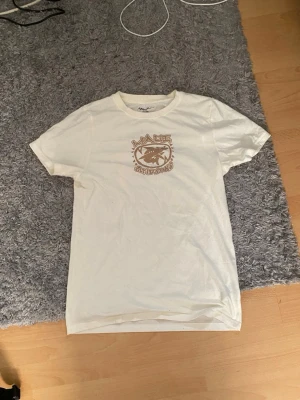 Vit t-shirt med hajtryck maui and sons - Vit t-shirt med brunt och turkost hajmotiv både fram och bak. T-shirten har rund hals och korta ärmar. Trycket har en retro känsla med grafiska detaljer. Perfekt för dig som gillar unika prints och streetwear vibes.