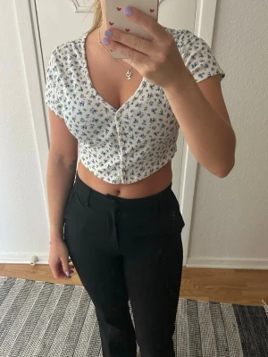 Vit croppad blommig topp - Söt croppad topp i vitt med blått blommönster och v-ringning. Toppen har korta ärmar, knappar framtill och är figurnära i modellen. Perfekt för varma dagar och ger en fräsch sommarkänsla.