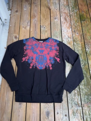 Marcello Burlon svart tröja - Cool svart sweatshirt med ett stort färgstarkt tryck av ett lejon i rött och blått över bröstet. Tröjan har rund halsringning, långa ärmar och ribbade muddar vid ärmslut och nederkant. Perfekt för dig som vill sticka ut med en unik design.