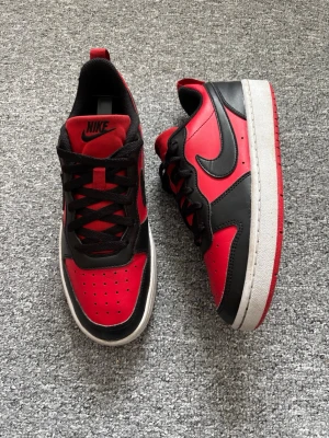 Nike svart-röda låga sneakers - Snygga låga sneakers från Nike i svart och rött med klassisk swoosh på sidan. Skorna har svart snörning, röd plös med Nike-logga och vit sula med röd undersida. Tillverkade i läder och textil för en sportig look. Varan är använd och är lite sliten och smutsig.