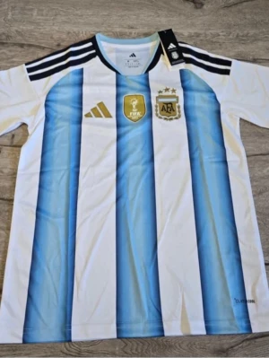 Argentina fotbollströja Adidas - Säljer en klassisk Argentina fotbollströja från Adidas med blå och vita vertikala ränder, svarta detaljer på axlarna och officiella klubbmärken på bröstet. Tillverkad i lätt och ventilerande material, perfekt för träning eller match.