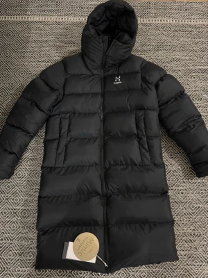 Svart lång dunjacka från Haglöfs - Snygg svart lång dunjacka från Haglöfs med huva och två stora framfickor. Jackan har en quiltad design med breda paneler och dragkedja framtill. Perfekt för kalla vinterdagar och riktigt varm tack vare dunfyllningen. Retail price 299 euros.