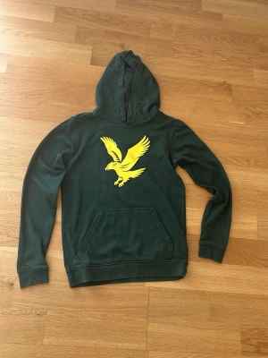 Grön hoodie med gul örn Lyle & Scott - Mörkgrön hoodie från Lyle & Scott med stor gul örn på bröstet. Klassisk känguruficka framtill och huva. Tillverkad i mjukt bomullsmaterial som är skönt att ha på sig. Perfekt för dig som gillar streetwear och vill sticka ut med en snygg logga.