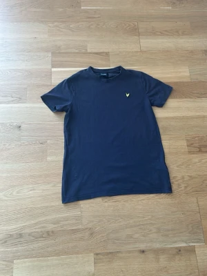 Mörkblå lyle&scott t-shirt  - En stilren mörkblå t-shirt i bomull med korta ärmar och rund hals. På bröstet sitter en liten broderad gul logga som ger en snygg detalj. Perfekt till jeans eller shorts för en enkel och clean look.