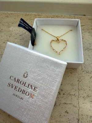 Halsband med hjärthänge i guldton - Modell: Baguette Heart Necklace  Färg: Crystal  Material: 18k guldplätering på mässing  Stenar: Swarowski kristaller  Längd: 45-50cm justerbart  Helt ny.