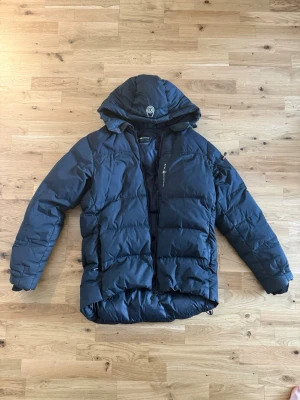 Sail racing vinter jacka - Snygg helsvart dunjacka med stor, justerbar huva och dragkedja framtill. Jackan är perfekt för kalla vinterdagar. Materialet är tåligt och håller värmen bra. Den ända defekten är ett litet hål som jag sytt igen på vänster ärm. Priser kan diskuteras vid snabb affär.