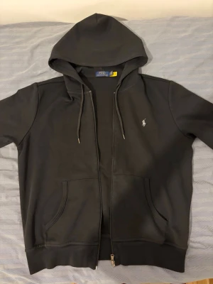 Polo Ralph Lauren Hoodie - Svart hoodie med dragkedja från Polo Ralph Lauren. Klassisk design med huva och dragsnören, två fickor framtill och den ikoniska broderade loggan på bröstet. Väldigt mjuk och skön känsla. Perfekt skick, Inga defekter alls på koftan. Storlek M. Pris kan diskuteras, Säljer även byxorna för 100 kr tillsammans och 120 separat.