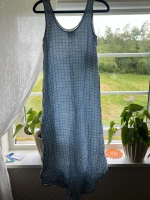 Blå långklänning med mönster - Luftig blå långklänning med diskret mönster och ärmlös design. Perfekt för varma dagar med sitt tunna material och avslappnade passform. Klänningen har rund halsringning och faller snyggt längs kroppen.
