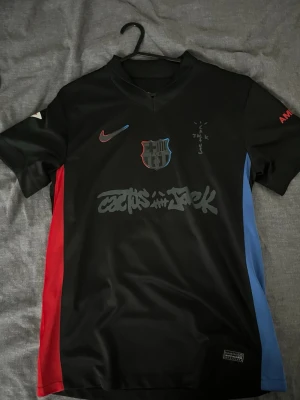 Barca X Travis Scott - Snygg svart FC Barcelona matchtröja från Nike med röda och blå detaljer på sidorna. Tryck med klubbmärke, Nike-logga och grafiskt motiv på bröstet. På ryggen står det 'Lamine Yamal' och nummer 19 i rött. Tillverkad i ett lätt och ventilerande material.